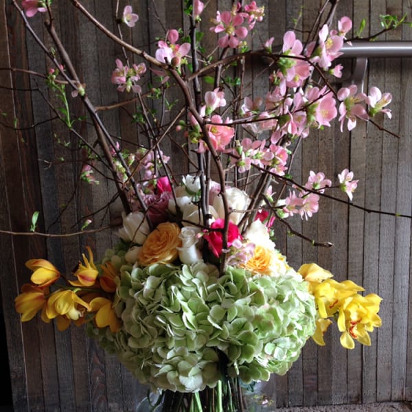 Tall floral arrangement with pink blossoms, hydrangeas, and yellow flowers in a glass vase