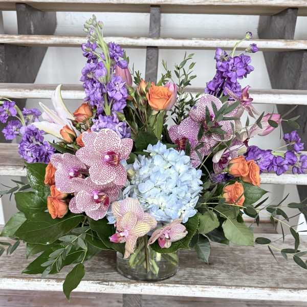 Mixed bouquet with purple blooms, orange roses, blue hydrangea, and orchids in a glass vase