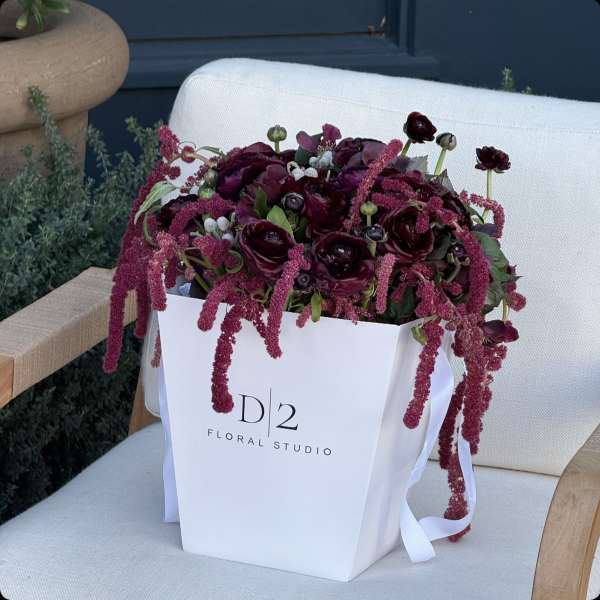Deep burgundy floral arrangement in a white gift bag with ribbon handles
