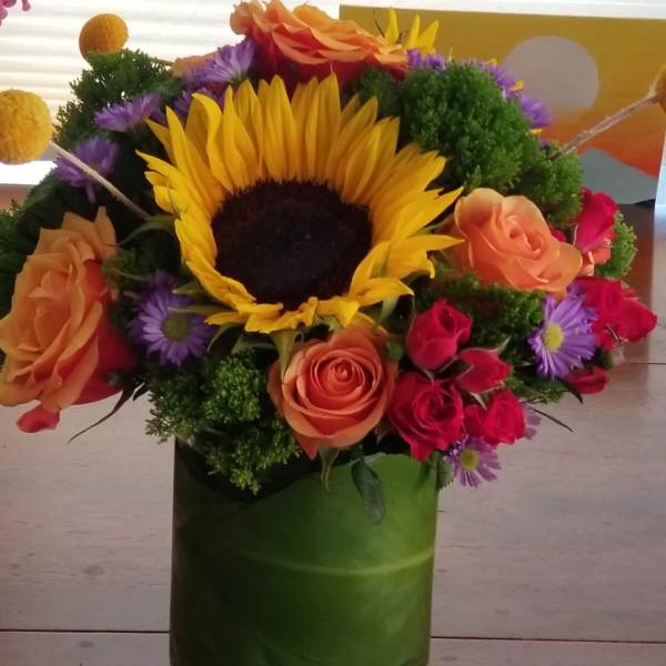 Bright sunflower, orange and red roses, and purple blooms arranged in a leaf-wrapped glass vase
