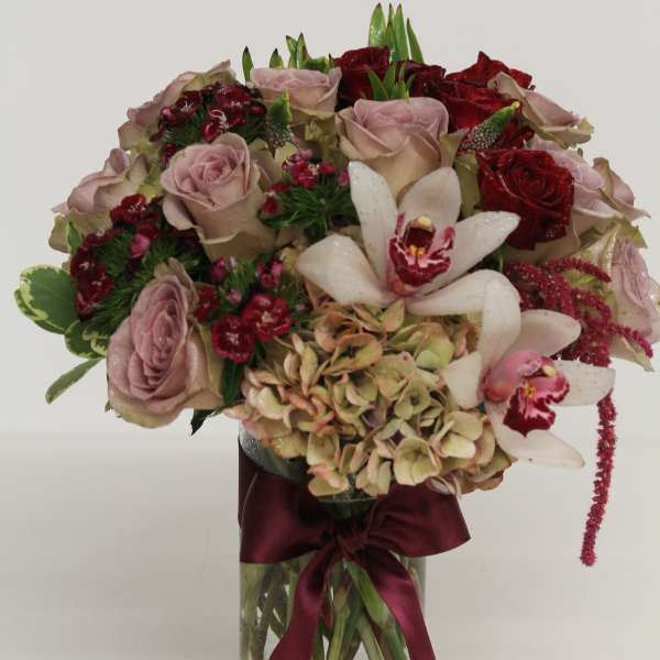 Bouquet of dusty pink roses, red roses, orchids, and hydrangea in a glass vase with a burgundy ribbon.