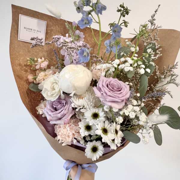 Pastel bouquet of lavender roses, white daisies, and mixed blooms wrapped in brown paper with a purple ribbon.