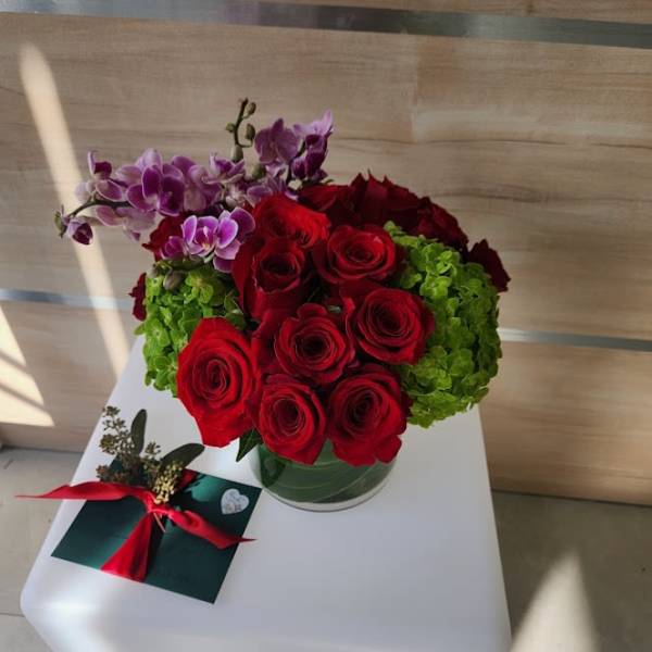 Low glass vase of red roses with green hydrangeas and purple orchids beside a ribbon-tied card
