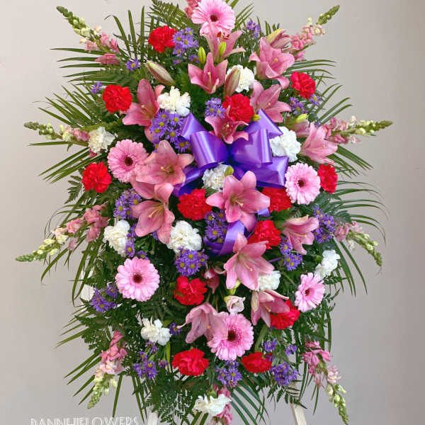 Standing floral spray with pink lilies, gerberas, carnations, and purple ribbon