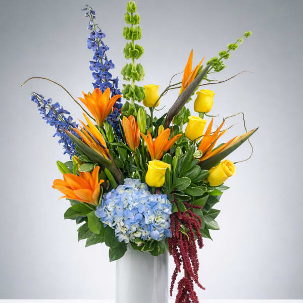 Tall mixed bouquet in a white vase with orange lilies, yellow roses, and blue flowers