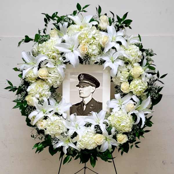 Large white floral wreath with lilies and roses on an easel framing a black-and-white portrait