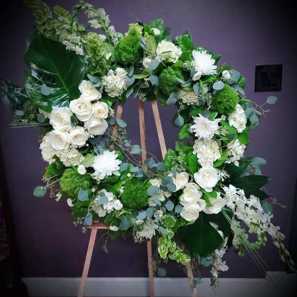 Large white and green standing wreath with roses, mums, orchids, and large glossy green leaves on a wooden easel.