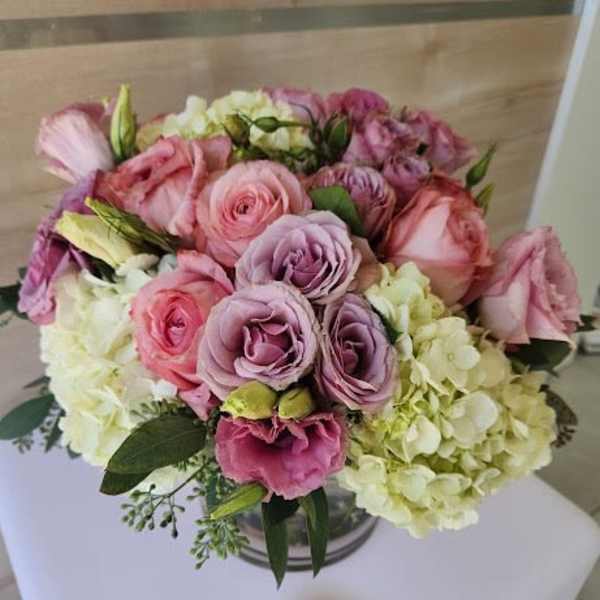 Low bouquet of pink roses, lavender lisianthus, and white hydrangeas in a clear glass vase