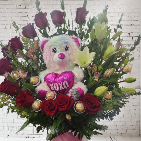 Red roses and lilies arranged with a teddy bear holding a pink heart.