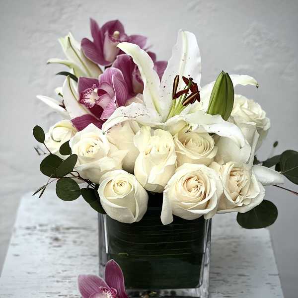White roses and lilies arranged in a square glass vase with purple orchids