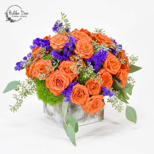 Orange roses with purple filler flowers in a square silver vase