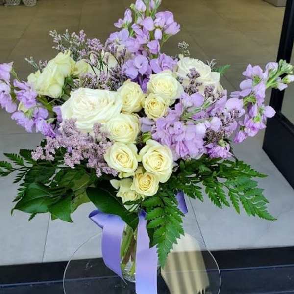 Vase arrangement of cream roses and light purple flowers with ferns and a lavender ribbon