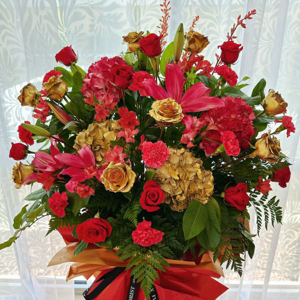 Tall standing arrangement of red roses, lilies, carnations and gold-toned blooms wrapped in red and gold paper.