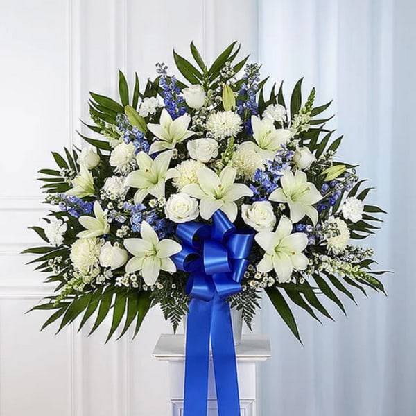 White lilies and roses with blue flowers and a ribbon on a stand