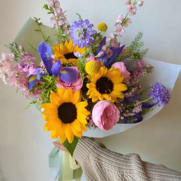 Handheld bouquet with sunflowers, pink roses, and purple blooms wrapped in paper