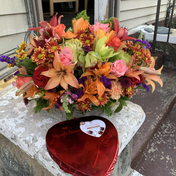 Bright mixed bouquet with roses, lilies, and chrysanthemums beside a heart-shaped chocolate box.