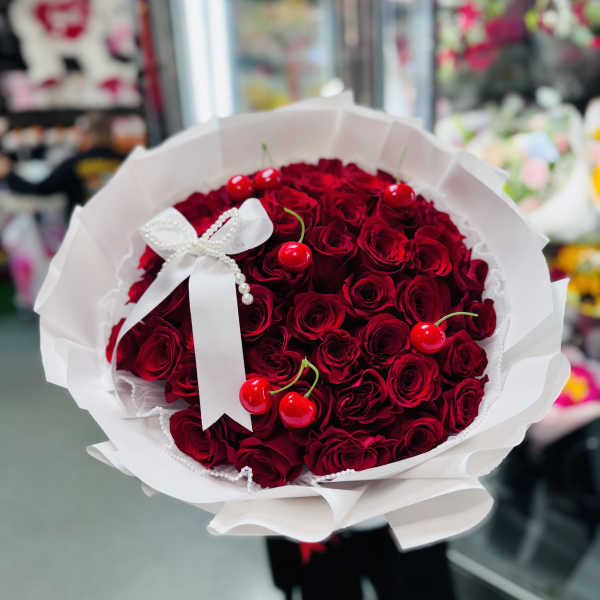 Round bouquet of deep red roses with faux cherries and a white bow in white paper wrapping