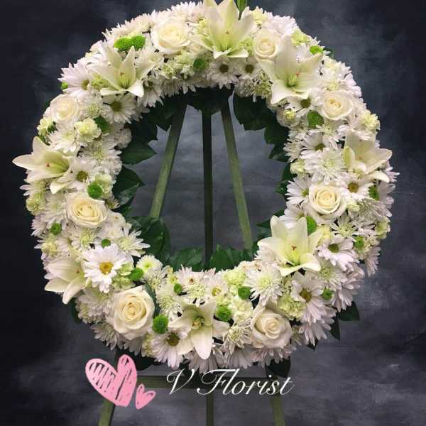 Large white and green standing wreath with roses, lilies, and daisies on a tripod stand.