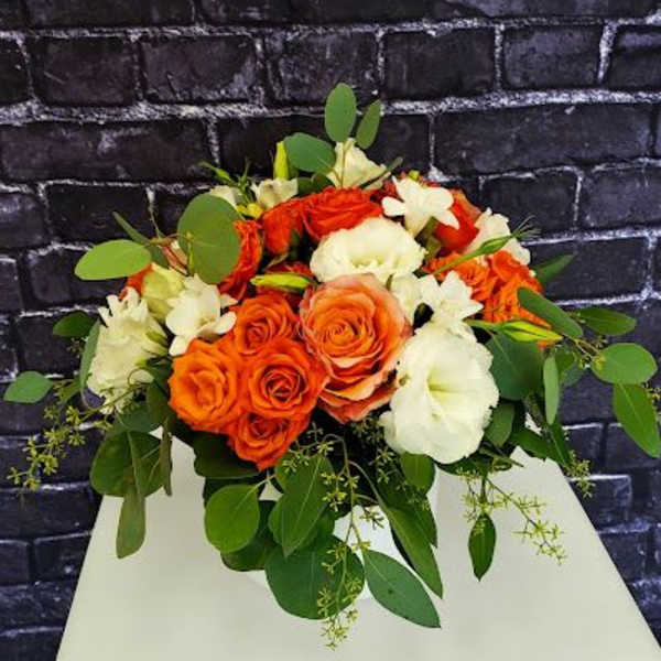 Low arrangement of orange roses and white blooms on a white pedestal with a dark brick backdrop