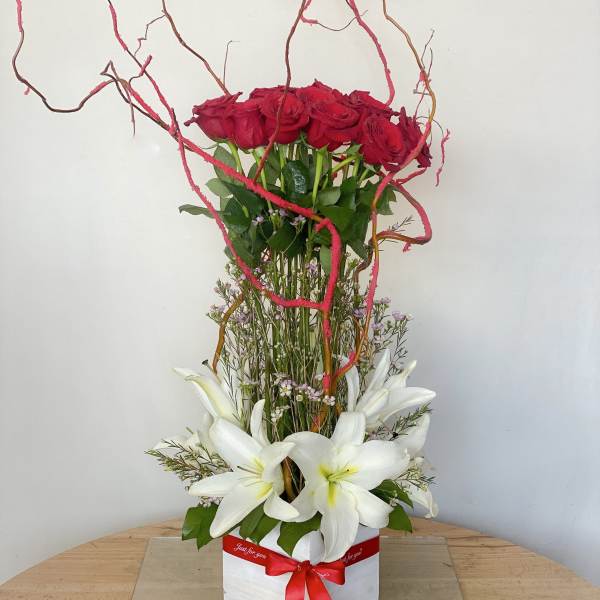 Tall arrangement of red roses and white lilies in a white box with a red ribbon