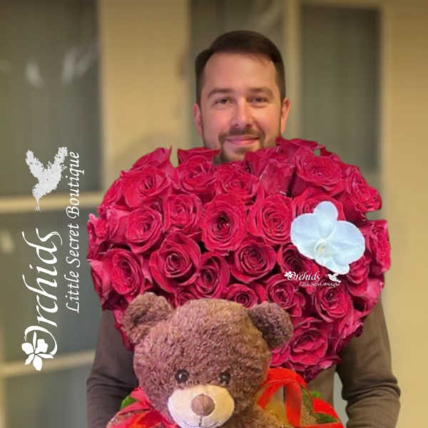 Man holding a heart-shaped rose arrangement with a teddy bear and ribbon