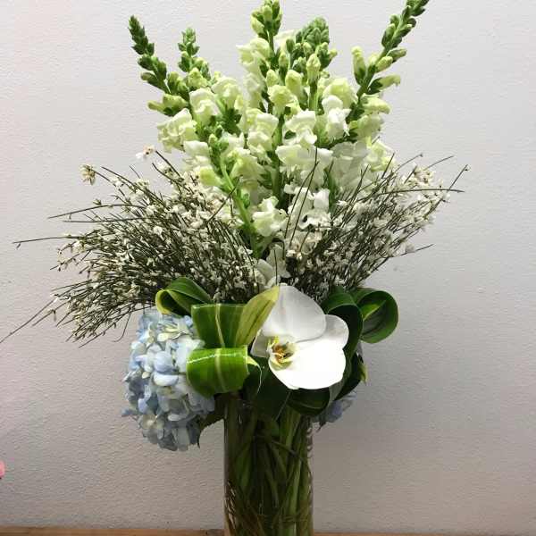 Tall white and green floral arrangement in a glass vase with blue hydrangea and orchid