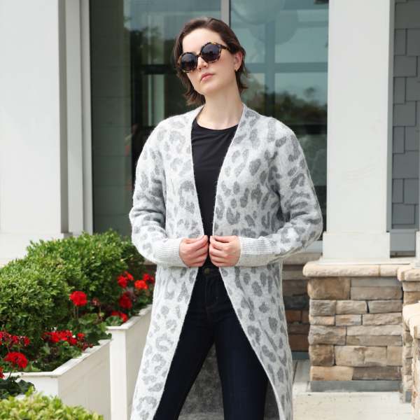 Woman in a long gray patterned cardigan and dark jeans standing by white planters with red flowers