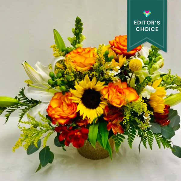 Mixed bouquet with orange roses, sunflowers, white lilies, and red blooms in a woven container