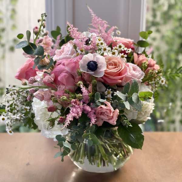 Pink and white mixed bouquet with roses and anemones in a round clear glass vase on a wooden surface.