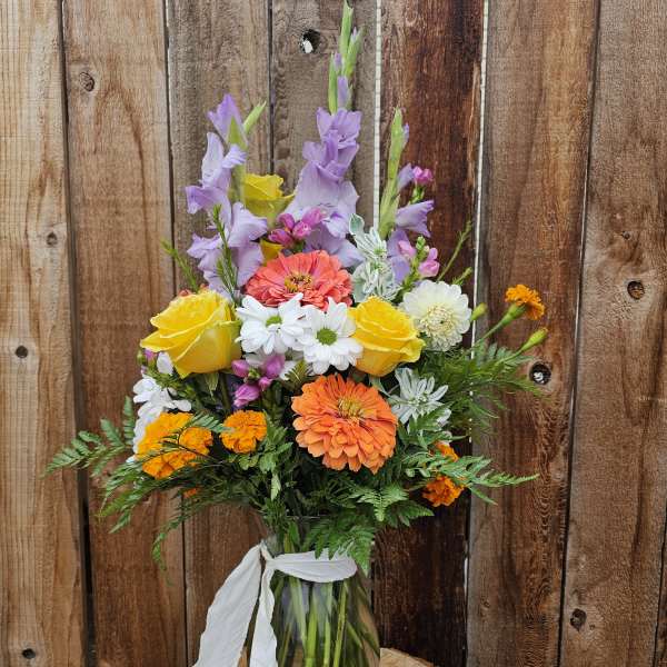 Tall mixed bouquet of orange, yellow, and purple flowers in a clear glass vase with white ribbon.