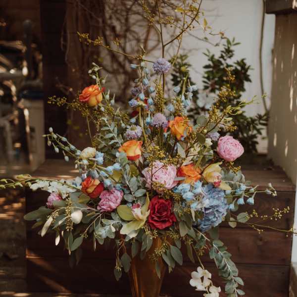 Tall floral arrangement in a gold vase with pink, orange, blue, and white blooms