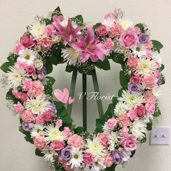 Heart-shaped standing wreath with pink lilies, roses, carnations, and white daisies on a green easel.
