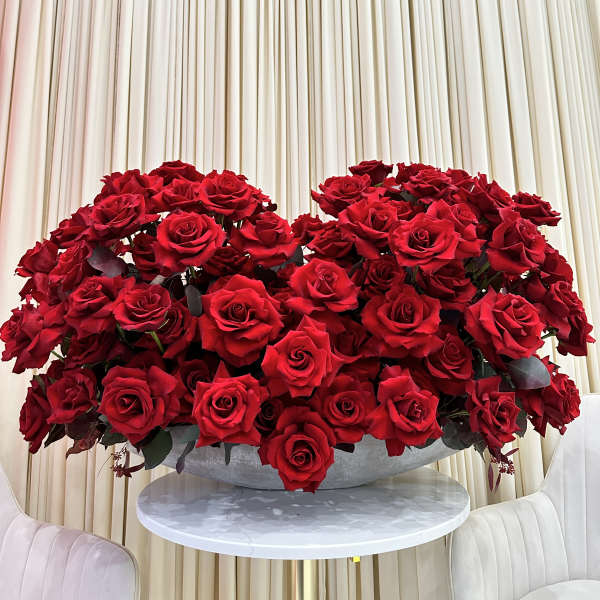 Large low arrangement of dozens of red roses in a wide bowl on a white table with cream drapery backdrop