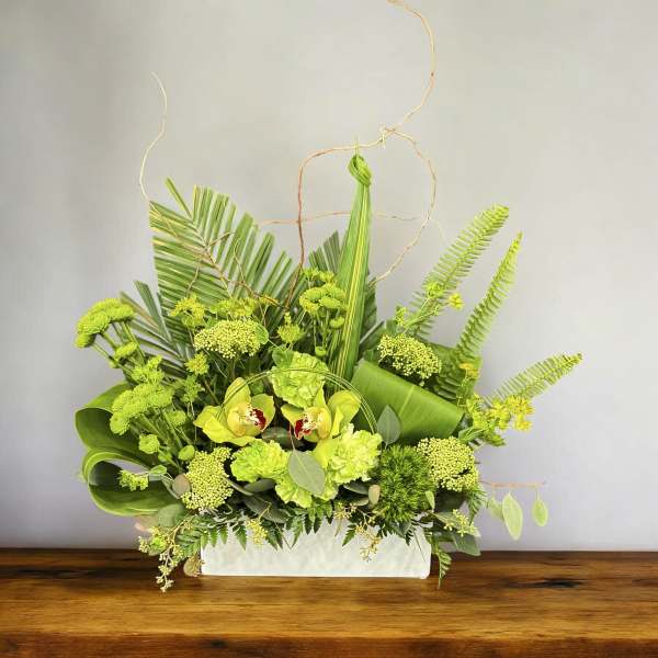 Green floral arrangement in a white rectangular container with orchids and tropical foliage