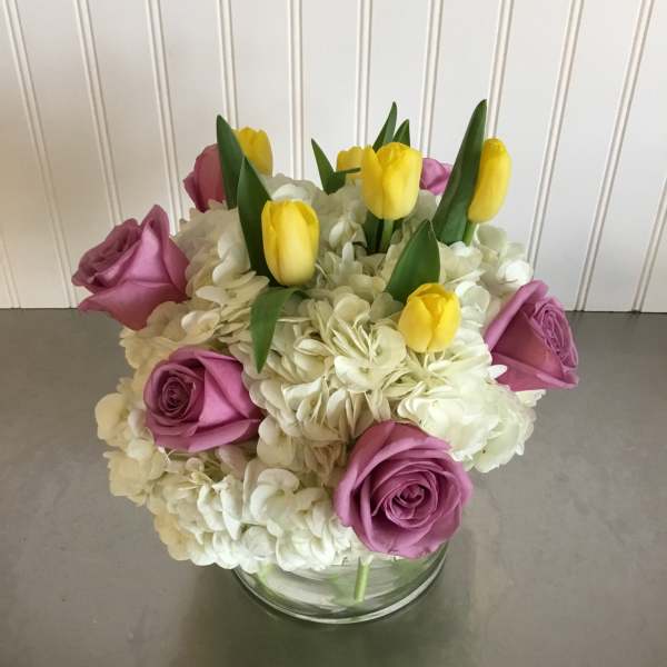 Compact arrangement of yellow tulips, lavender roses, and white hydrangeas in a clear glass vase
