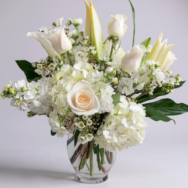 White floral arrangement in a clear glass vase with roses, calla lilies, and hydrangeas