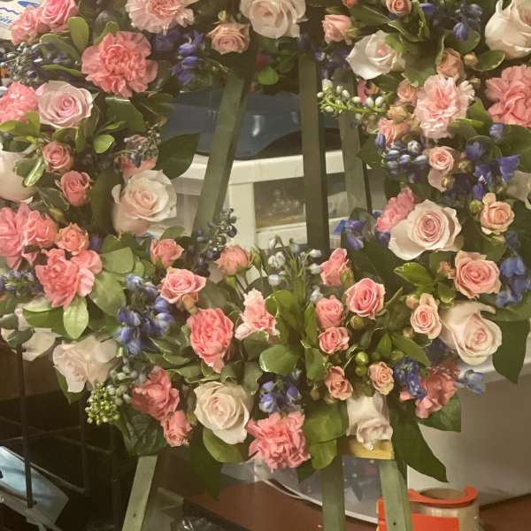 Standing wreath of pink roses and carnations with blue accent flowers on a green easel.