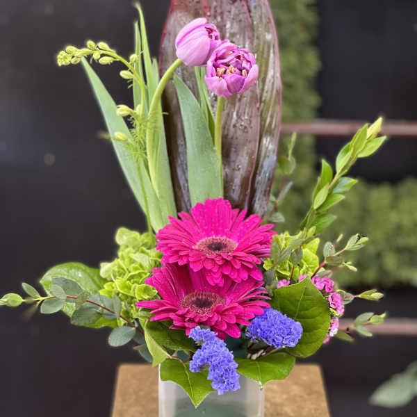 Pink gerbera daisies and tulips in a square vase with mixed greenery