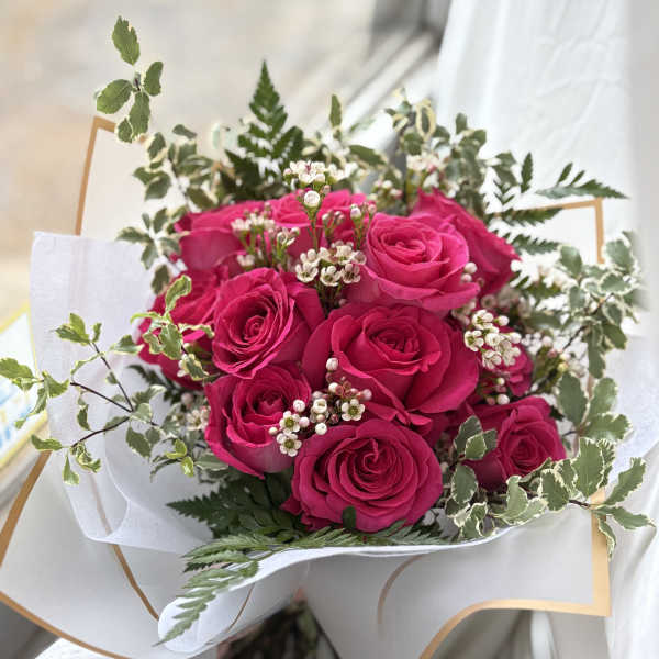 Hand-tied bouquet of hot pink roses with small white filler flowers wrapped in white paper.