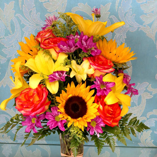 Bright mixed bouquet in a clear glass vase with sunflowers, roses, and purple daisies