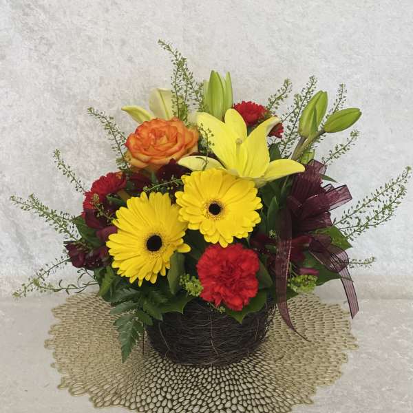 Mixed bouquet with yellow gerbera daisies, lilies, and red carnations in a dark basket