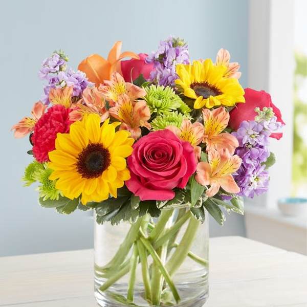 Mixed bouquet with sunflowers, roses, and purple flowers in a glass vase