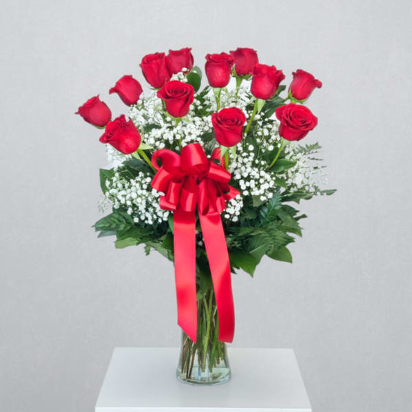 Dozen red roses with white filler flowers in a clear vase with a red ribbon bow