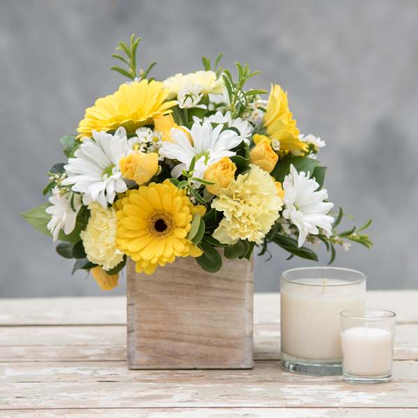 Yellow and white flower arrangement in a wooden box beside two candles