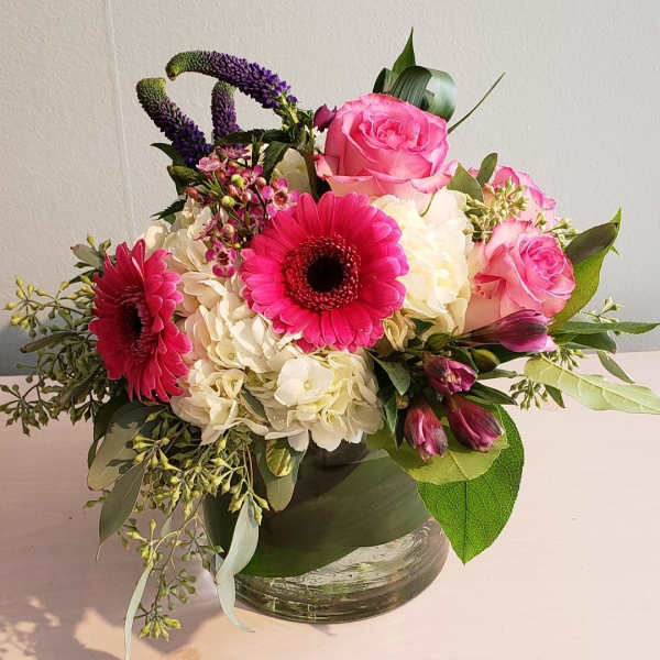 Low round arrangement of pink gerbera daisies, pink roses, and white hydrangeas in a clear glass vase