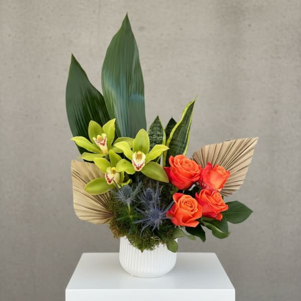 Modern arrangement with green orchids, orange roses, palm fans and large green leaves in a white vase.