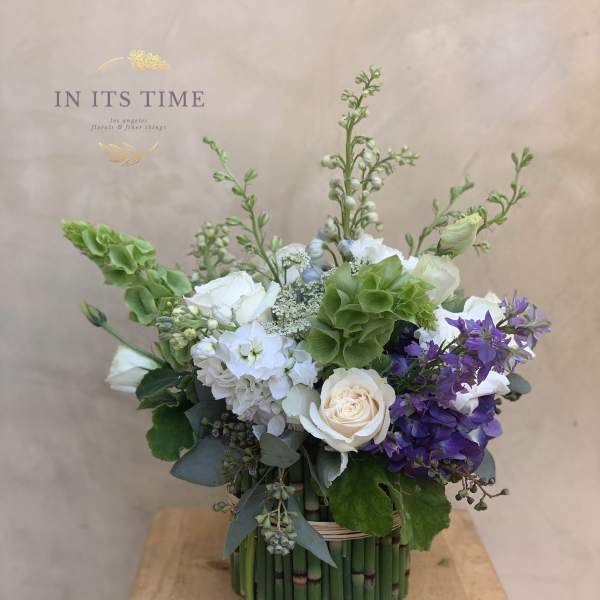 Mixed bouquet of white, green, and purple flowers in a bamboo-wrapped container