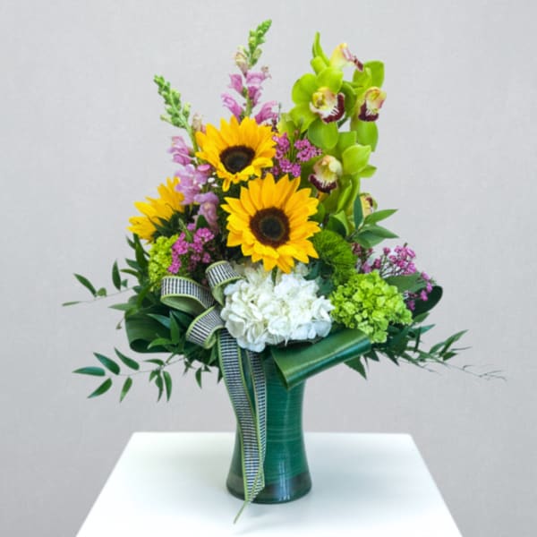 Tall mixed arrangement with sunflowers, green orchids, hydrangeas, and pink blooms in a leaf-wrapped vase.
