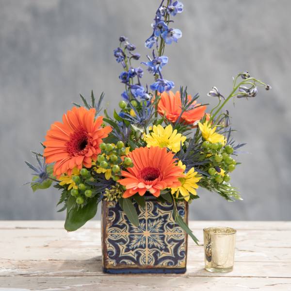 Orange and yellow flowers in a patterned square vase with a small candle holder beside it