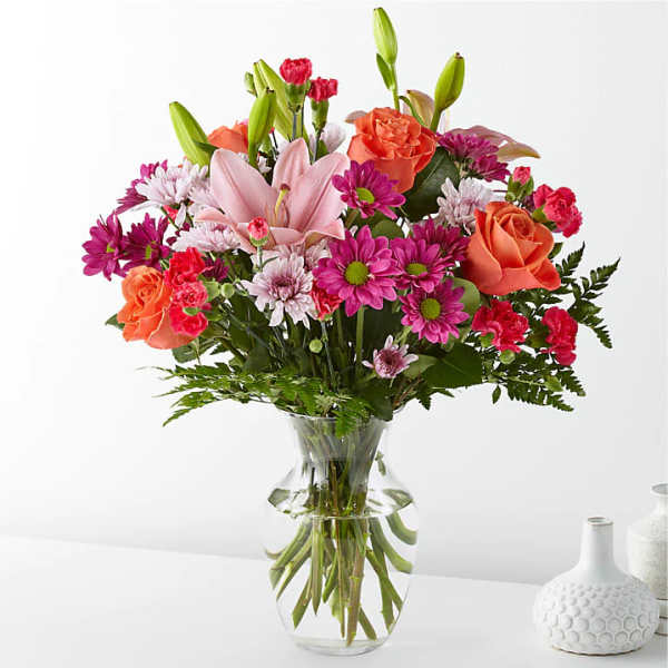 Mixed bouquet of pink lilies, orange roses, and magenta daisies in a glass vase